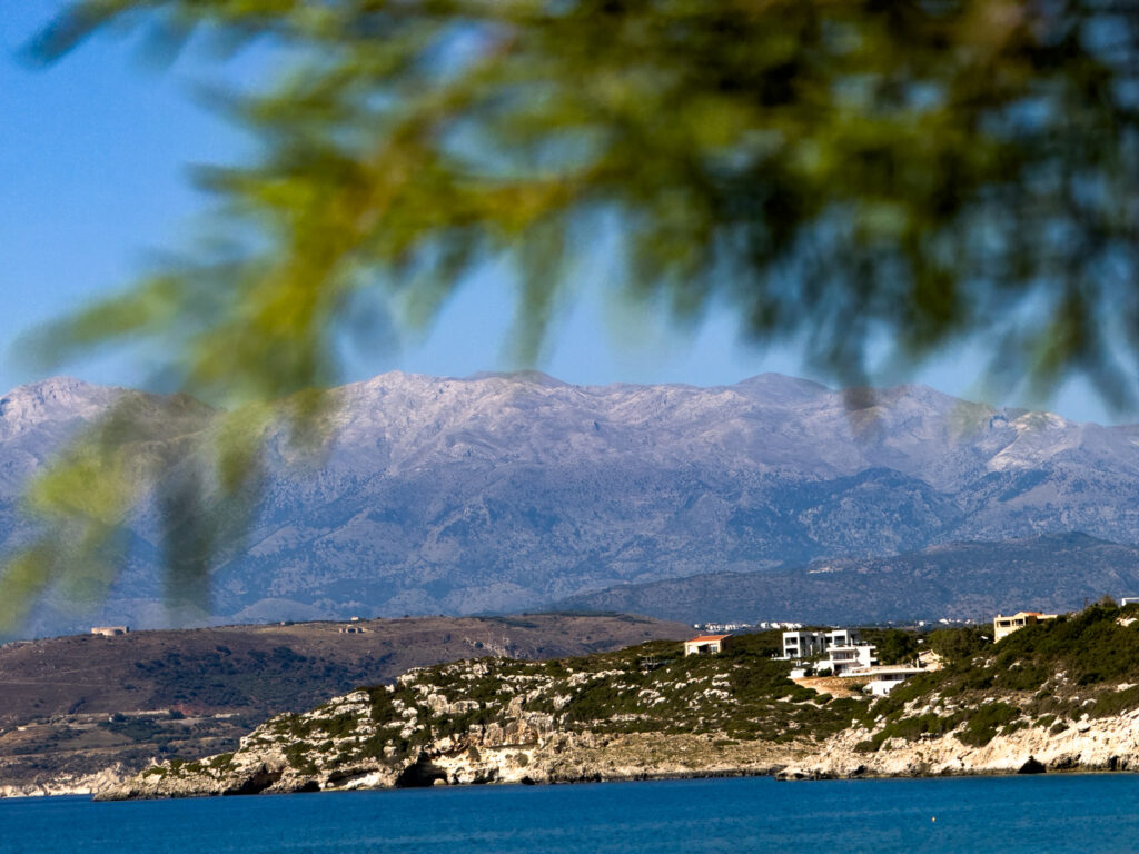 Marathi Beach in Crete with calm turquoise water in the foreground and a rocky shoreline lined with white hillside villas. Rugged mountains stretch across the background under a clear blue sky, while soft green branches frame the top of the scene. The image captures the peaceful coastal landscape and natural beauty of Marathi Beach.