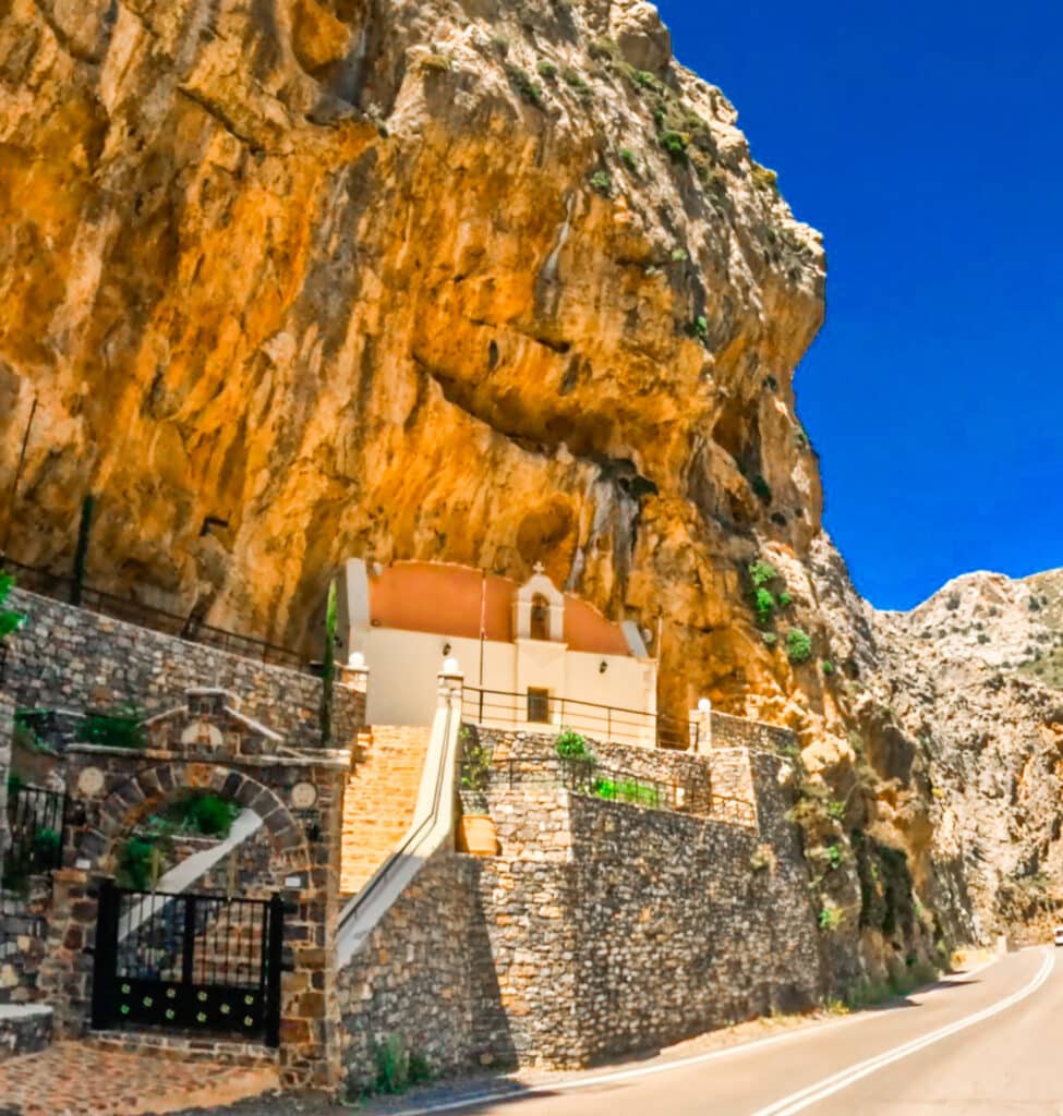 Stone-built monastery nestled against the vivid orange cliffs of Kourtaliotiko Canyon in Crete, Greece, with a bright blue sky above and a winding path leading to the entrance. The dramatic contrast between the rugged rock face and the architecture highlights the secluded, spiritual setting.