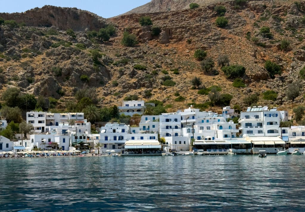 A group of white buildings next to a body of water. View of Loutro village from the water on a private Crete sailboat tour