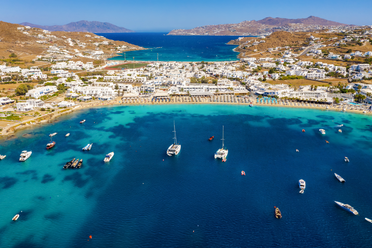 a city on the water Aerial view to the popuar bay of Ornos on the island of Mykonos, Greece, with many yachts moored in front of the beautiful beach