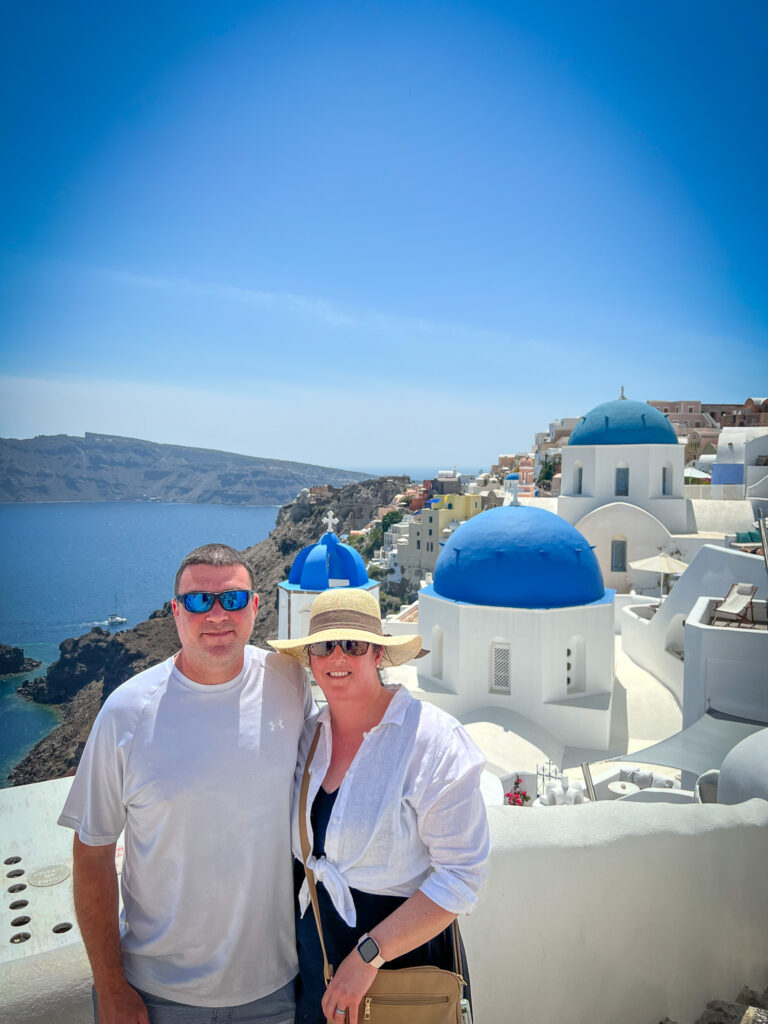 a man and woman posing for a picture in front of a blue and white building 3 domes of Santorini Oia