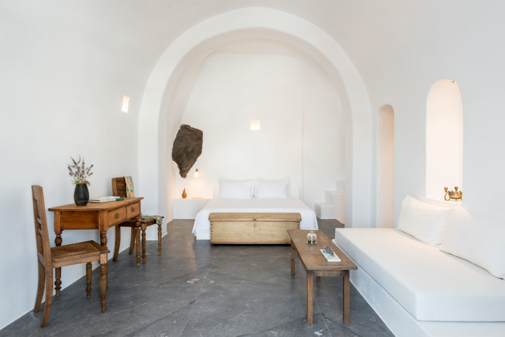 Minimalist cave-style suite at Chelidonia Luxury Suites in Oia Santorini, featuring a vaulted ceiling, cozy white bedding, rustic wooden furniture, and soft natural lighting ideal for a honeymoon stay.