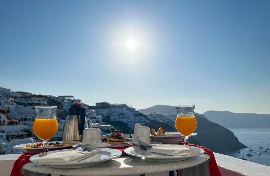 Romantic breakfast setup for two at Chelidonia Luxury Suites in Oia, with fresh juice, pastries, and a caldera view under the morning sun—perfect for a Santorini honeymoon moment.