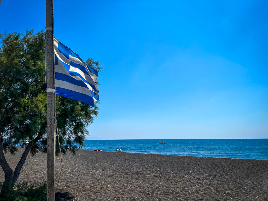 A Greek flag flutters beside a windswept tree on a black sand beach in Santorini, with the deep blue Aegean Sea stretching to the horizon under a clear sky. The dark volcanic sand contrasts with the bright blue water, highlighting the island's unique landscape.