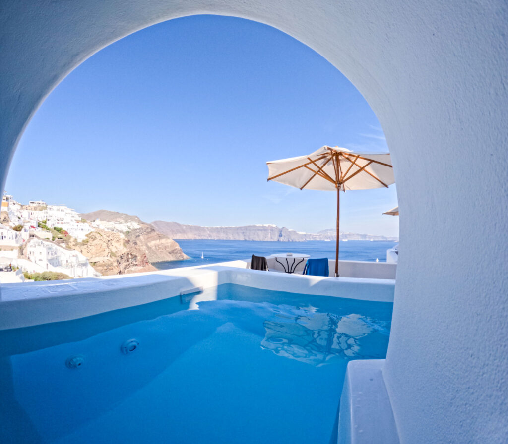 Private plunge pool at Chelidonia Luxury Suites in Oia Santorini, framed by a white arch with a view of the caldera and cliffs under a sunlit sky. A shaded patio with an umbrella and seating creates a serene setting for a honeymoon escape.