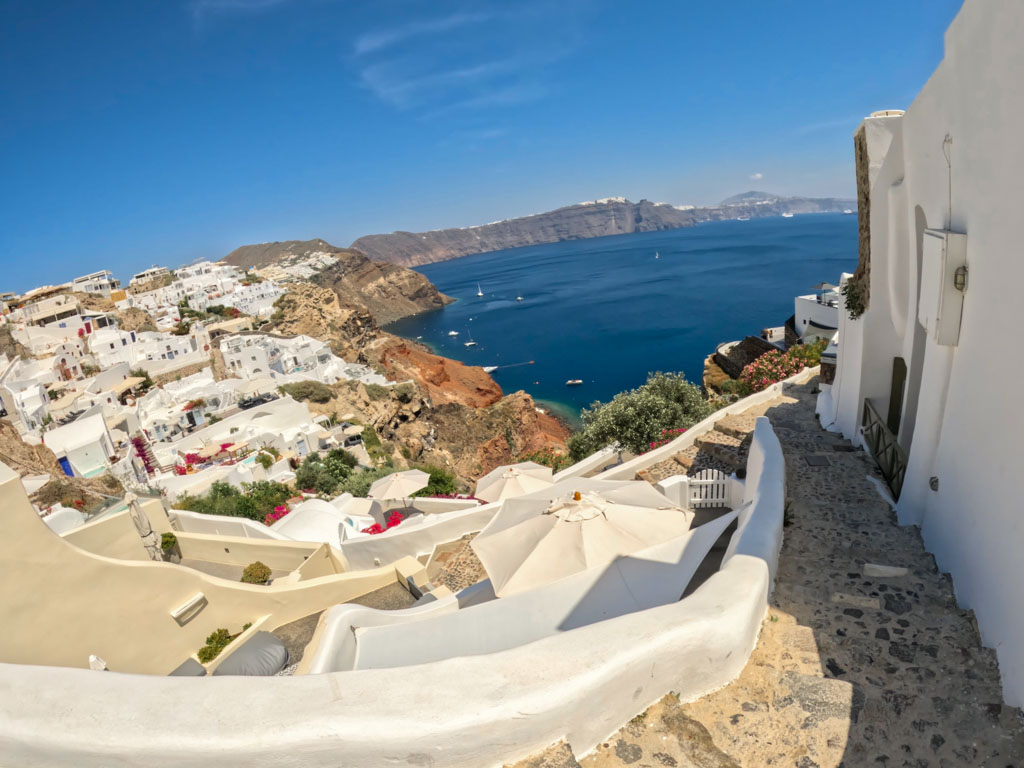 Peaceful, sunlit view of Santorini’s whitewashed buildings cascading down a cliffside toward the deep blue Aegean Sea, seen from a quiet stone path. The calm setting reflects why travelers often ask, “is Santorini safe” for a serene getaway.