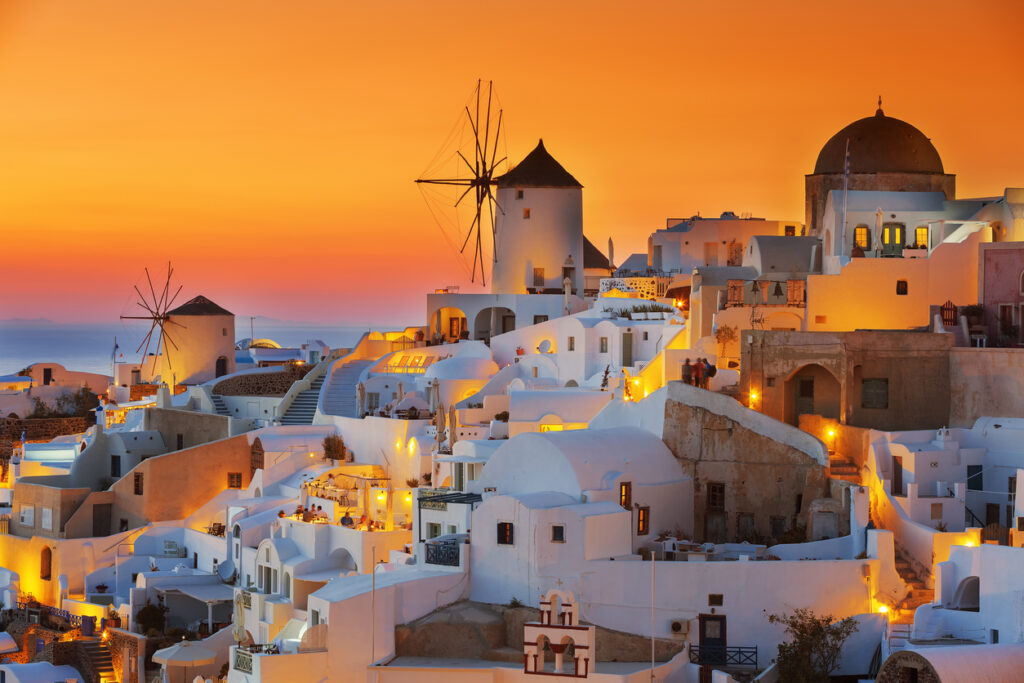 Iconic windmills and whitewashed buildings of Oia, Santorini glow under a vivid orange and pink sunset sky. Warm lights illuminate the cliffside village, capturing the magic of a classic Santorini sunset.