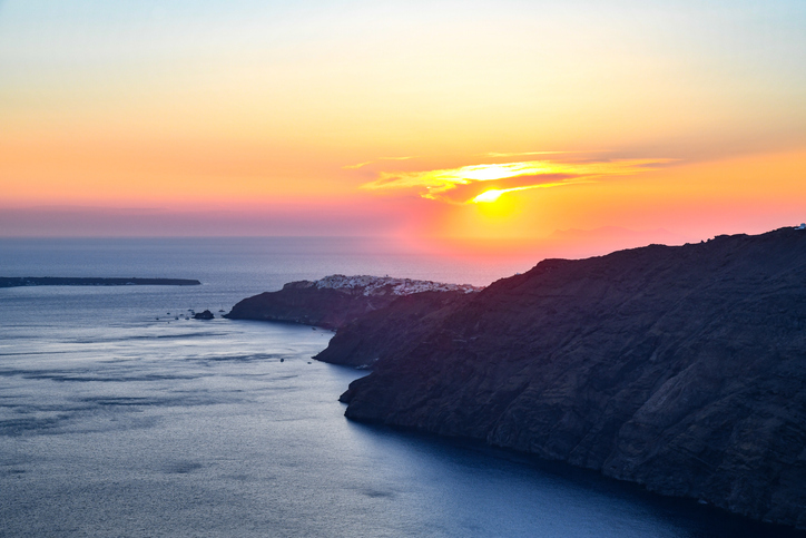 A glowing Santorini sunset over the Aegean Sea, with golden and pink hues reflecting on the water and silhouetted cliffs stretching into the distance. The peaceful view captures the dramatic coastline and soft evening light.
