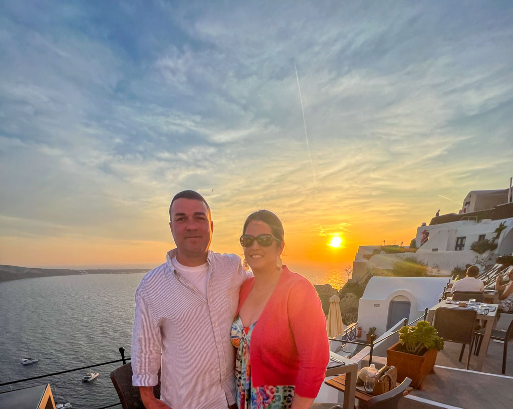 Couple posing at sunset on a terrace overlooking the sea at Ombra restaurant in Santorini. The golden sun sets over the water, casting a warm glow on the white-washed buildings and outdoor diners on the cliffside.