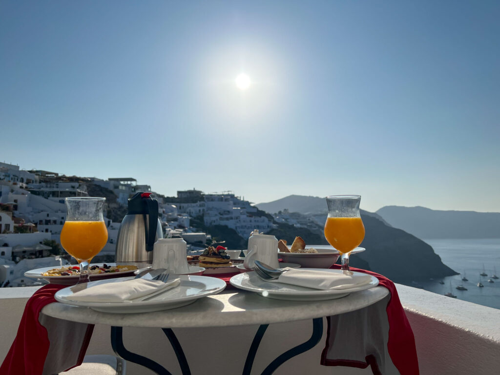Romantic breakfast setup for two at Chelidonia Luxury Suites in Oia, with fresh juice, pastries, and a caldera view under the morning sun—perfect for a Santorini honeymoon moment.