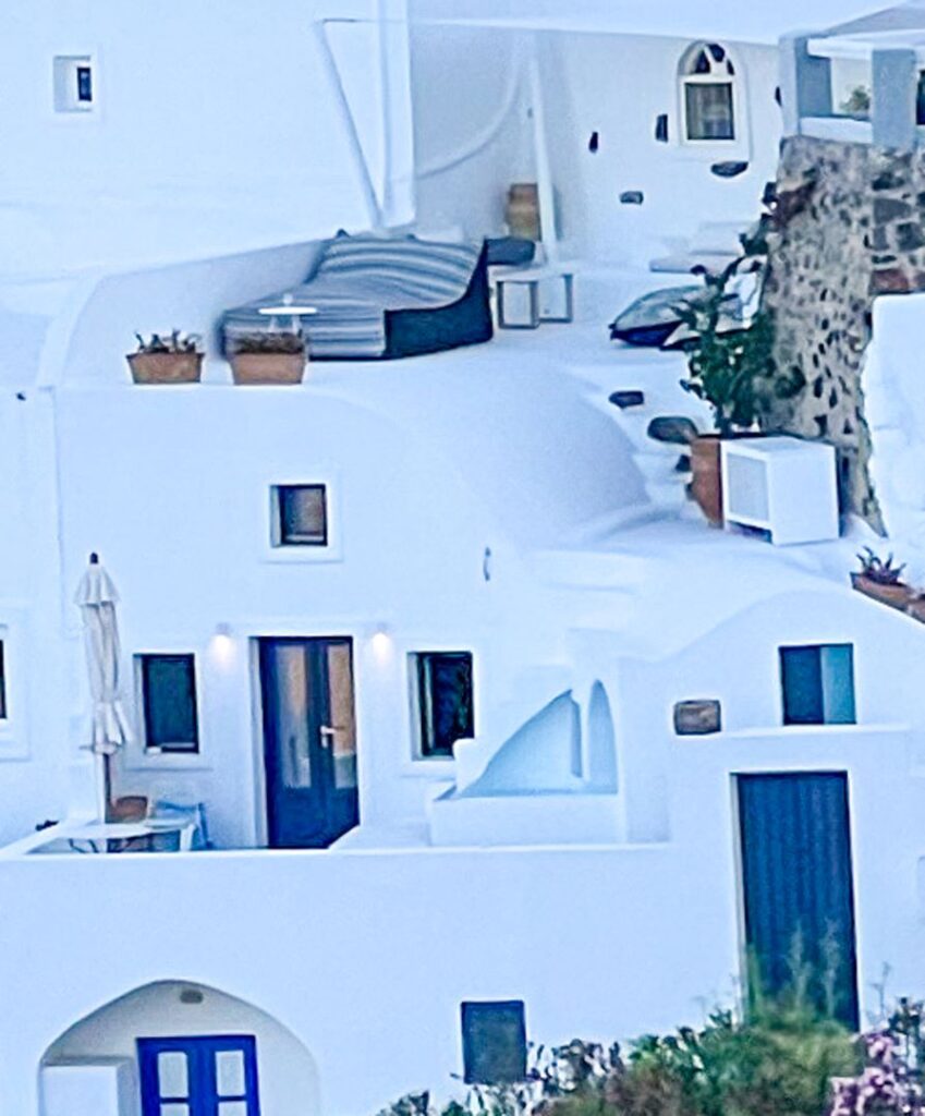 Close-up of a private terrace at Chelidonia Luxury Suites, showing curved white architecture, potted plants, and a shaded daybed overlooking Oia’s iconic scenery—capturing honeymoon serenity.