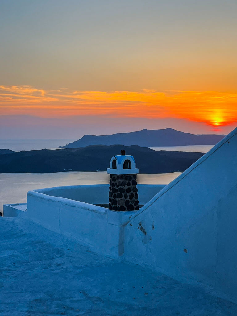 A vivid sunset over the sea in Fira, Santorini, with an orange and pink sky above dark volcanic islands, seen from a whitewashed terrace with a small stone chimney.