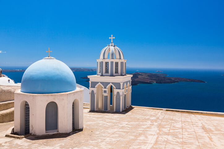 Two whitewashed church domes—one light blue and rounded, the other ornate with arches and topped by a cross—stand against a vivid blue sky overlooking the Aegean Sea. This is the Church of St. Mark the Evangelist in Firostefani, Santorini, with volcanic islands visible in the distance.