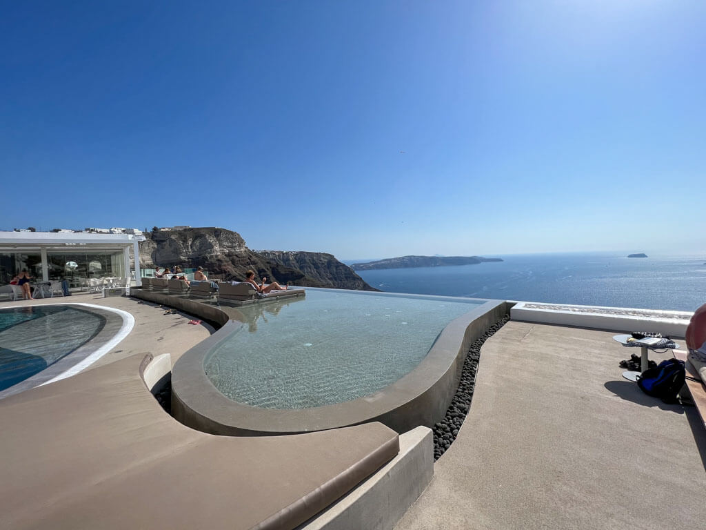 An infinity pool overlooking the caldera in Fira, Santorini, with people lounging on sunbeds and a sweeping view of the blue sea and cliffs under a clear sky.