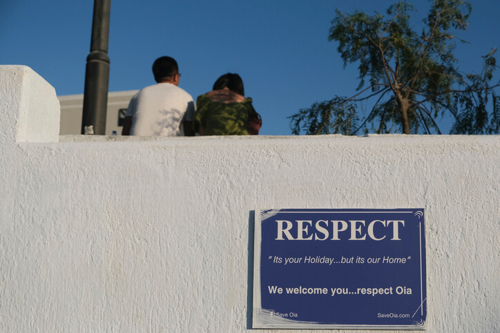 Two people sit on a whitewashed wall in Oia, Santorini, with a blue sign below them that reads, “RESPECT – It's your Holiday...but it's our Home. We welcome you... respect Oia.” A tree and lamppost are visible in the background under a clear blue sky.