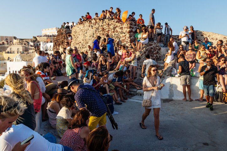 A large crowd of tourists gathers on the ruins of a stone castle in Oia, Santorini, waiting to watch the famous sunset. People are seated on steps and ledges, chatting and taking photos under the warm evening light.