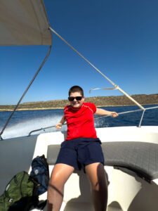 Boy wearing a red shirt and sunglasses relaxes on the back bench of a moving boat, holding onto the railing as water splashes behind him under a clear blue sky. A green backpack sits beside him, and dry, hilly coastline is visible in the background.