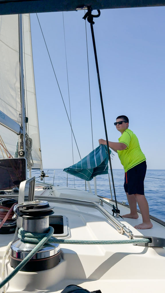 Boy in a neon yellow shirt and navy shorts stands barefoot on the deck of a sailboat, holding a folded striped beach towel near the mast under a clear blue sky. Calm sea surrounds the boat, with rigging and sailing equipment visible in the foreground.