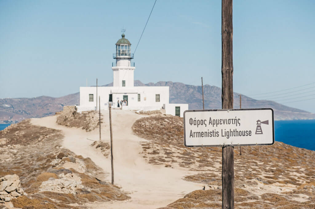 Remote view of the whitewashed Armenistis Lighthouse perched on a rocky hilltop in Mykonos, with a directional sign in Greek and English pointing the way.