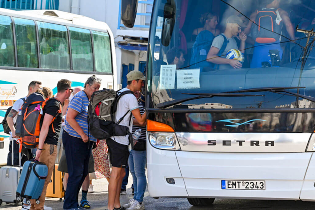 Tourists with backpacks and rolling luggage boarding a white Setra bus, heading toward a Mykonos port. A child is visible inside the bus near the front window.