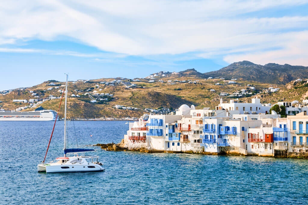 Whitewashed buildings with blue shutters perched at the water's edge in Mykonos’ Little Venice, with a sailboat drifting nearby and rolling hills in the background under a partly cloudy sky.