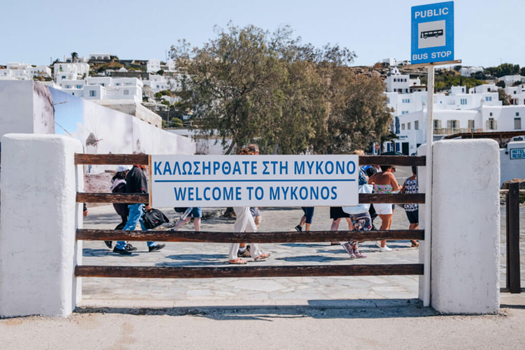 A whitewashed alleyway in Mykonos with a large sign that reads “WELCOME TO MYKONOS” in both Greek and English, with tourists walking past.
