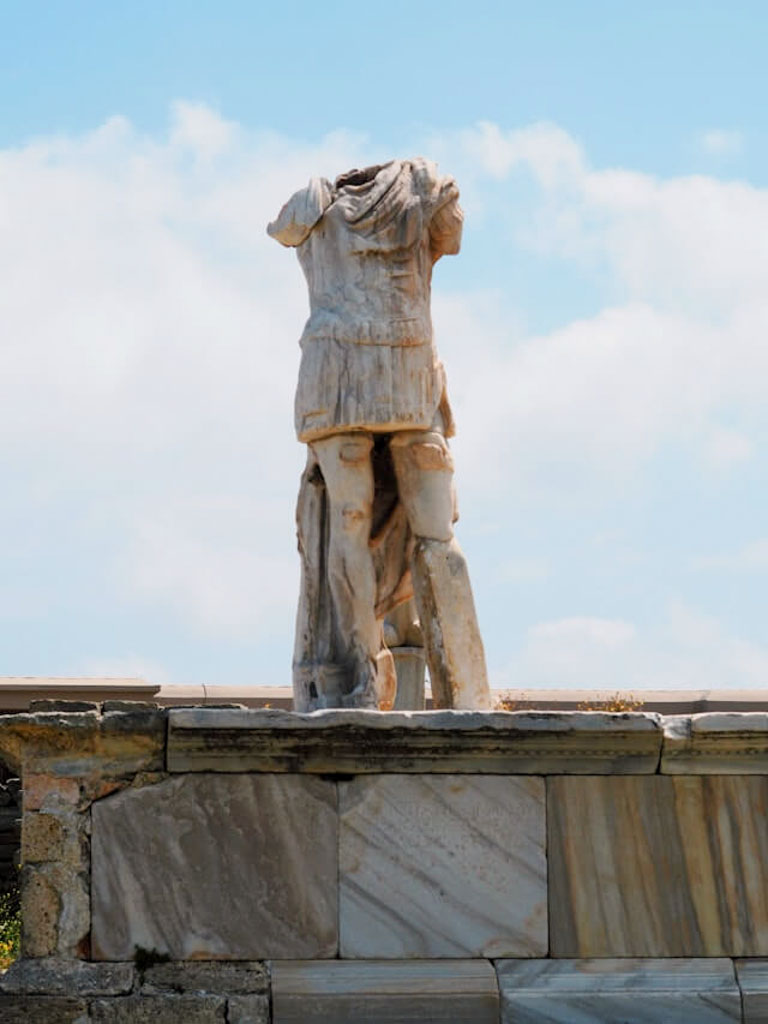 Headless marble statue of a standing male figure with detailed drapery, elevated on a stone platform under an open sky on Delos Island.