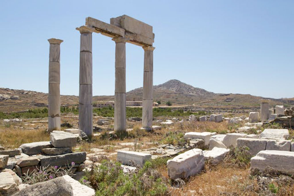 Ancient columns from a temple on Delos Island standing tall against a blue sky with distant hills and scattered stone fragments on the ground.