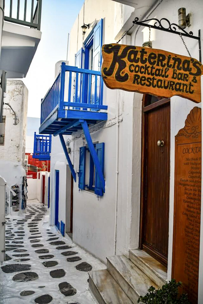 Narrow alley in Mykonos’ Little Venice with whitewashed buildings, blue shutters and balconies, and the wooden sign for Katerina's Cocktail Bar and Restaurant hanging above the entrance. The stone path curves through the charming Cycladic scene.