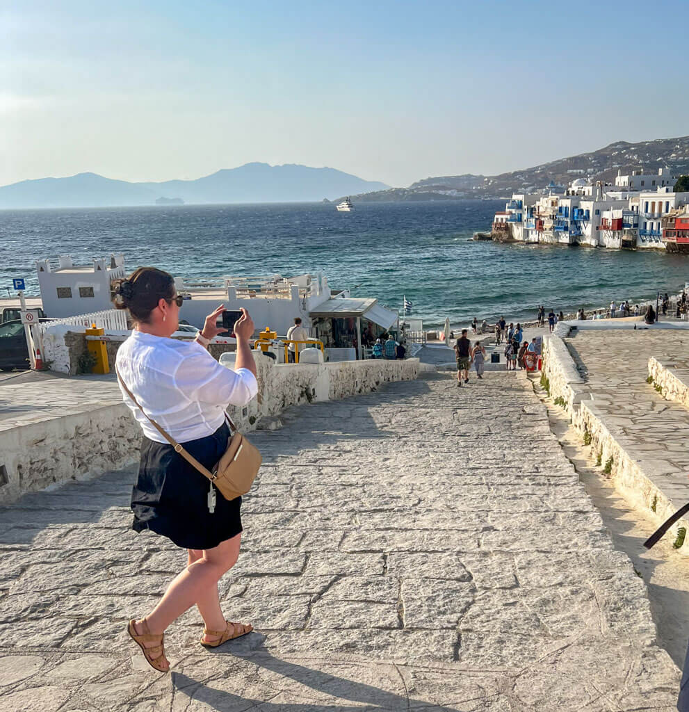 A woman in a white blouse and black skirt takes a photo while walking down a stone path toward the sea in Mykonos Little Venice. The scene captures colorful waterfront buildings, sunlit waves, and distant mountains, with tourists exploring the area below.