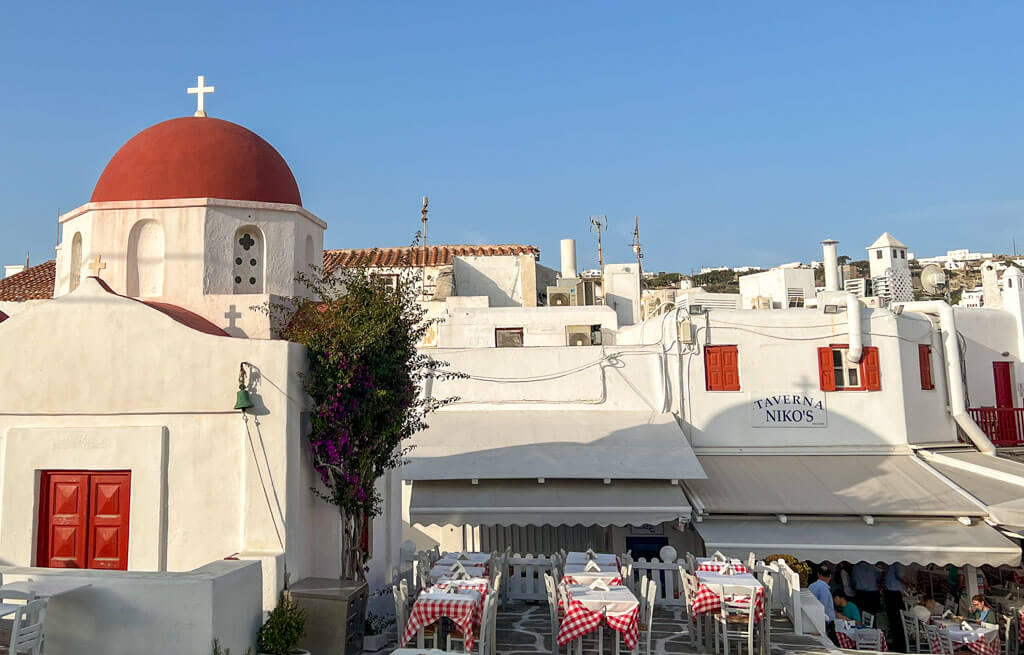 Close-up of the red-domed church and bustling Taverna Nikos in Mykonos. Tables line the stone courtyard, bathed in golden afternoon light.