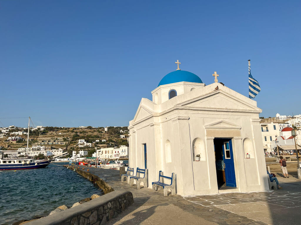 Iconic seaside chapel of Agios Nikolaos in Mykonos with a vibrant blue dome, Greek flag waving beside it, and moored boats in the background.