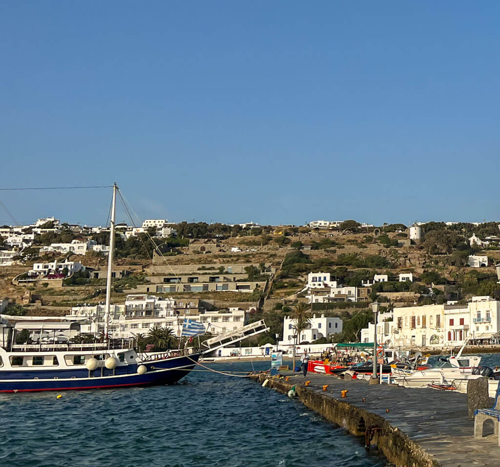 Pier in Mykonos lined with small fishing boats and a classic wooden sailboat. Hillside homes and white buildings dot the background under a clear blue sky.