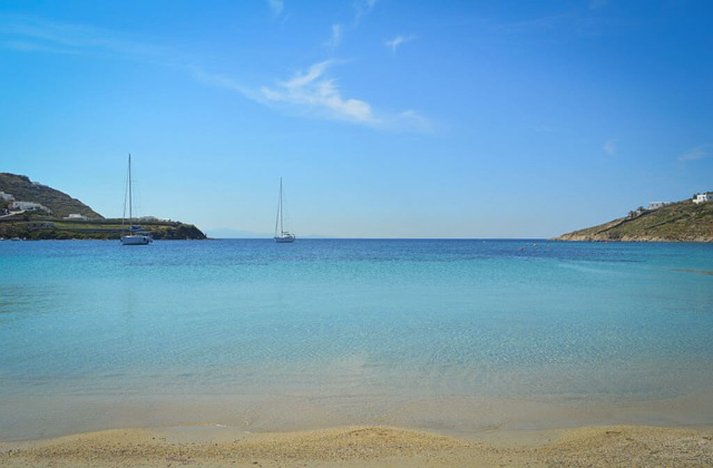 Calm shallow sea with a sailboat in the distance under a cloudless sky near Paradise Beach in Mykonos