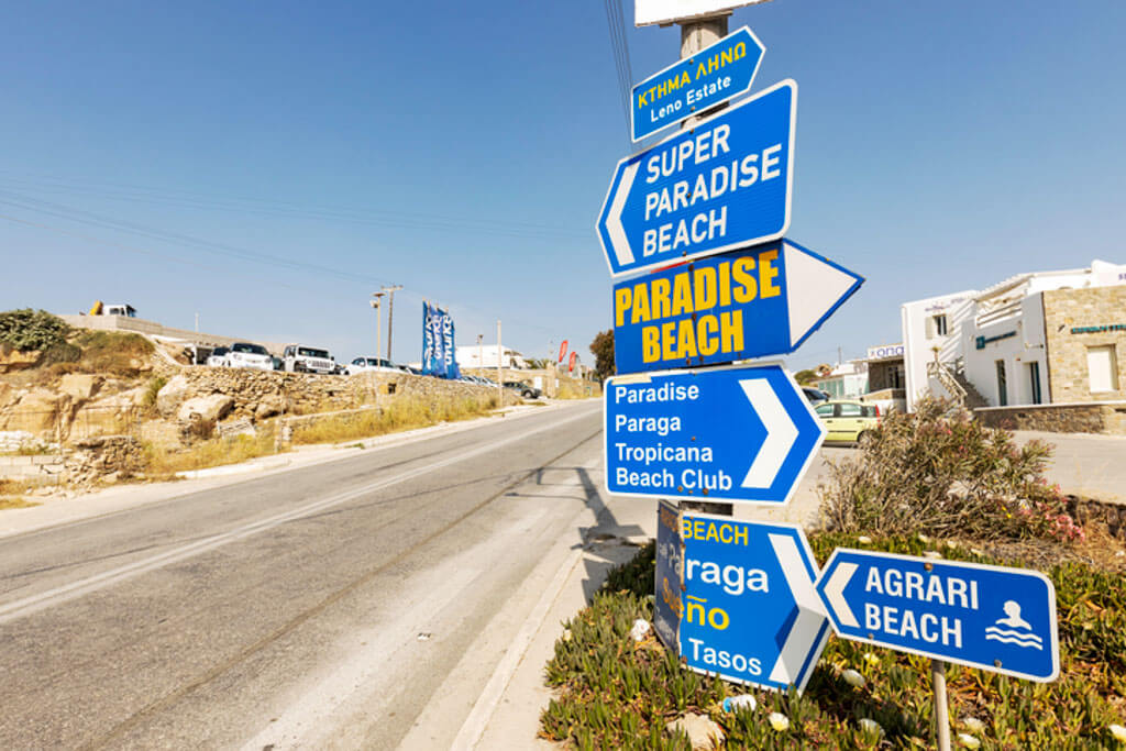 Cluster of blue road signs in Greek and English on a roadside in Mykonos, pointing toward popular beach destinations.
