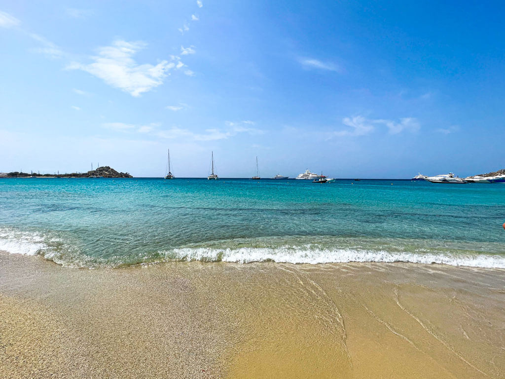 Shallow waves gently lapping onto the golden sand at Platis Gialos Beach in Mykonos, with turquoise water stretching out to sailboats anchored in the calm bay under a sunny blue sky.