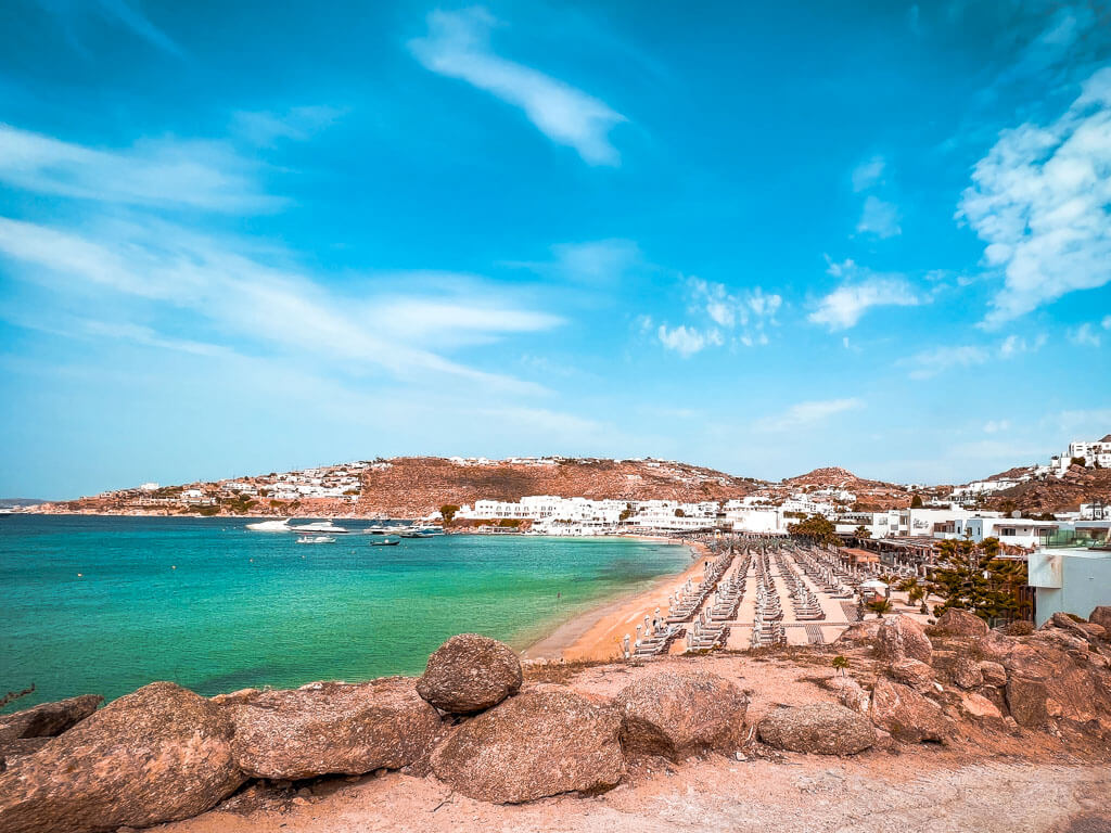 Bright turquoise waters and a sandy beach surrounded by rocky terrain on a sunny day in Mykonos. Platis Gialos Beach