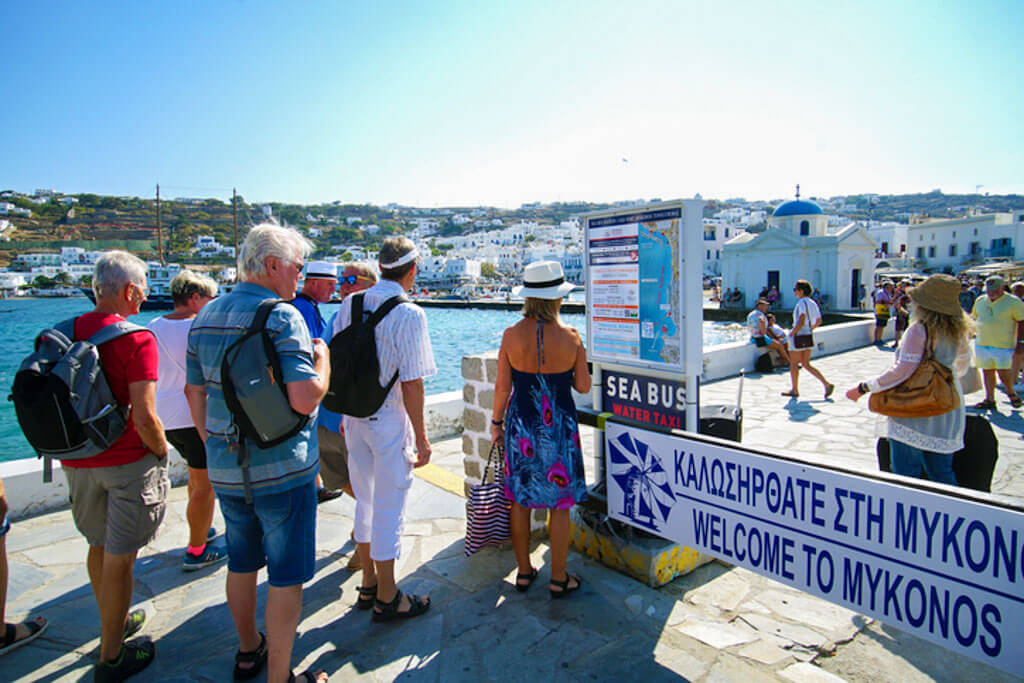 Tourists queue near a waterfront sign reading “Welcome to Mykonos” next to a Sea Bus water taxi map. The backdrop features white Cycladic buildings and a blue-domed chapel, capturing the charm of Mykonos Town.