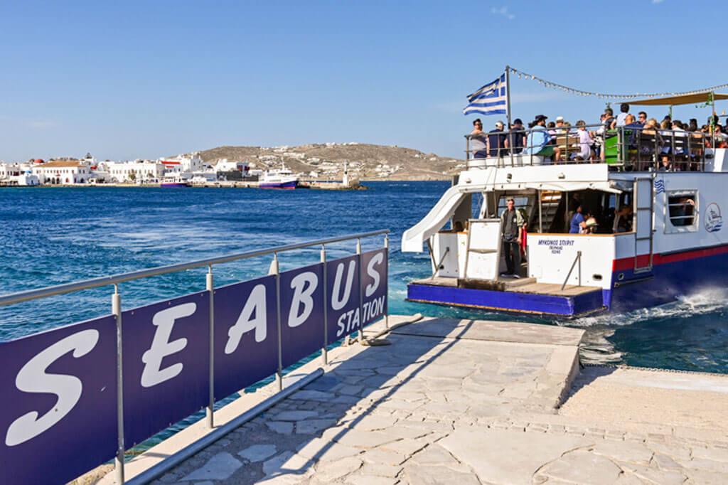 A ferry labeled “SEABUS” docked at a pier in Mykonos, with passengers on the upper deck enjoying ocean views under a Greek flag.