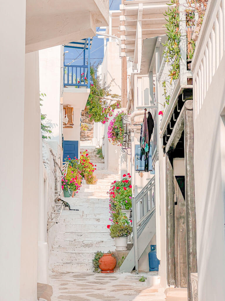 Charming alleyway in Mykonos with white stone steps and bright flowerpots lining the way. Pops of pink and red from blooming flowers add to the postcard-perfect setting.