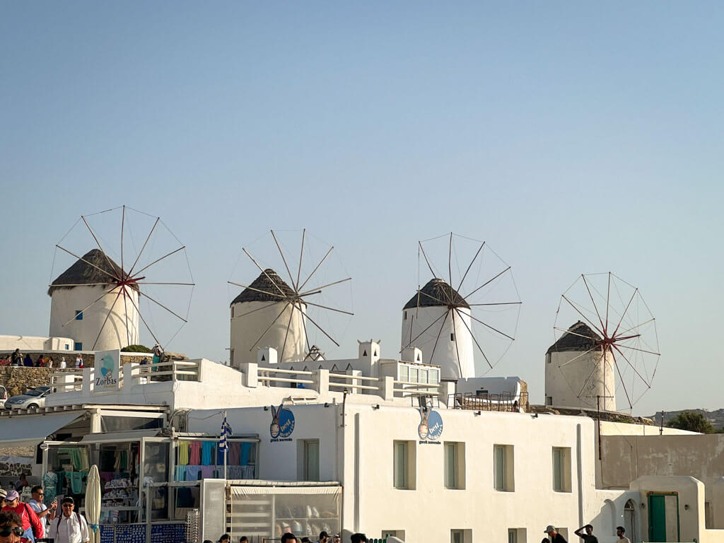 Five traditional white windmills with conical thatched roofs rise above a row of shops and whitewashed buildings in Mykonos, set against a clear blue sky. Tourists gather below, capturing one of the island’s most iconic views.