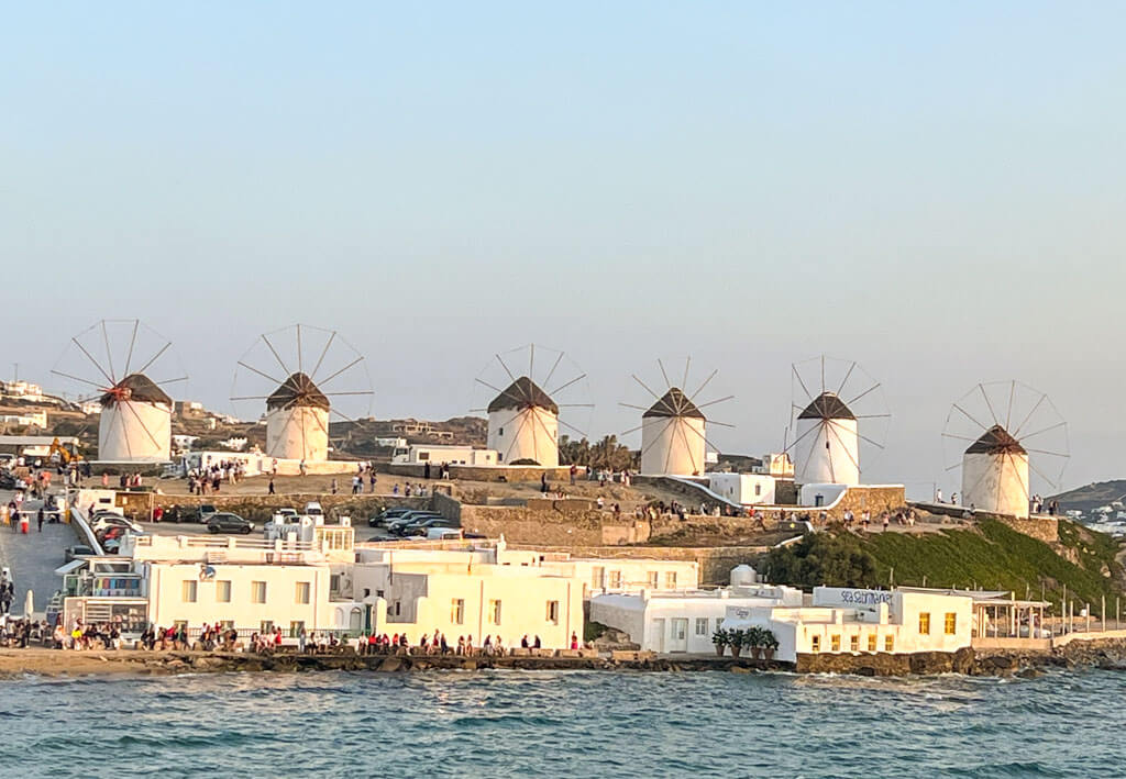 A row of iconic white windmills with thatched roofs stands tall on a hill above Mykonos town, overlooking whitewashed buildings and the shimmering sea at sunset.