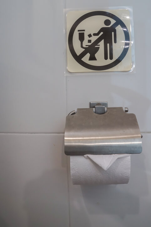 A bathroom wall in Greece with a metal toilet paper holder and a humorous sign above it showing a person throwing trash into a toilet with a line through it, indicating what not to do. The sign reflects common restroom etiquette reminders found in Greek public bathrooms.