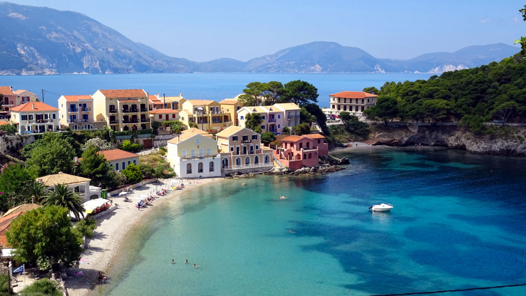 Colorful pastel houses with terracotta roofs line the curved shoreline of Assos in Kefalonia Greece overlooking a calm turquoise bay. A small pebble beach with sunbathers and swimmers sits beside clear shallow water while a lone boat floats near the center of the cove. The image captures the charm of this picturesque Ionian village framed by green hills and distant mountains.