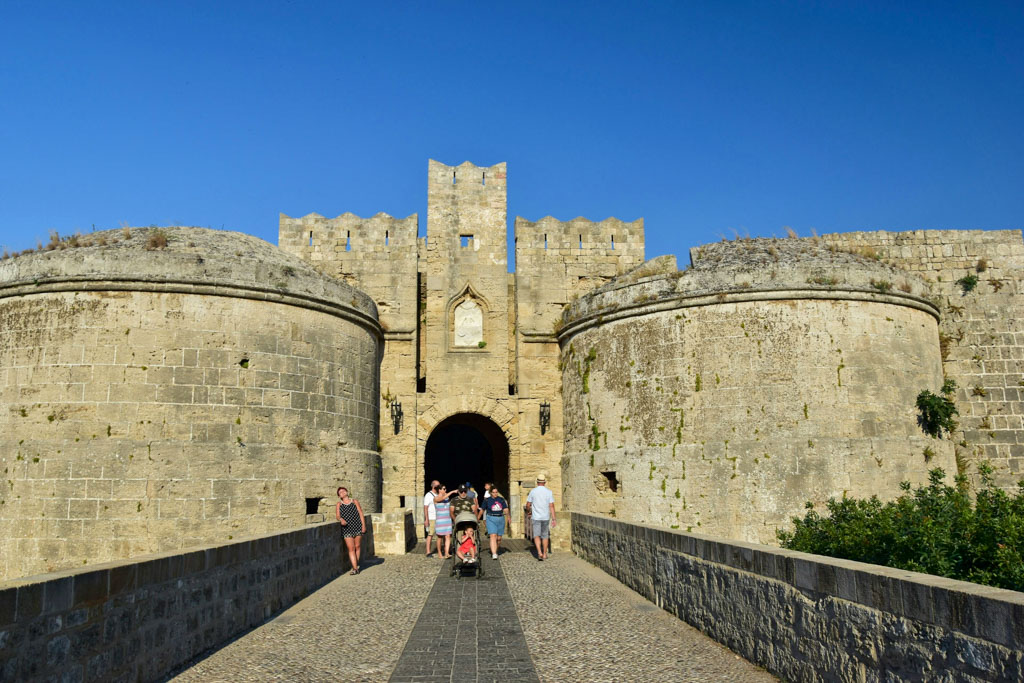 Stone bridge leading to the Gate D Amboise in Rhodes Greece framed by two massive round medieval towers and a tall central archway under a clear blue sky. Visitors walk toward the fortress entrance including a family with a stroller which highlights the scale of the historic stone walls. The image captures the grandeur and preserved architecture of this iconic Rhodes landmark.