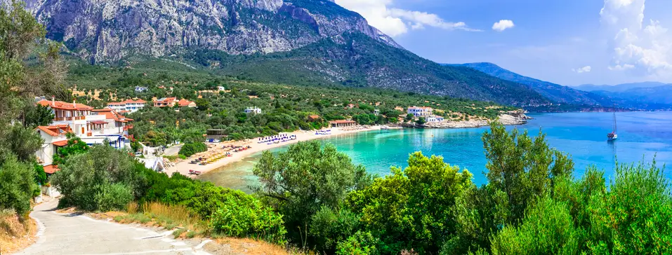 Turquoise water curves along Limnionas Bay in Samos framed by lush green trees and a sandy beach lined with sunbeds and umbrellas. White buildings with terracotta roofs sit at the base of a rugged mountain creating a peaceful coastal village setting. The image highlights the natural beauty and crystal clear sea that make Limnionas Bay a relaxing spot on Samos.