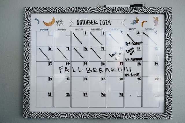 A wall mounted dry erase calendar labeled October 2024 with several dates crossed off in black marker. The words Fall Break are written across the middle, and notes on the right side include Buy jacket, Leave at 9am, VS. Hinesen, and VS. Pitt. The image shows a busy monthly schedule with school events and fall activities marked clearly.